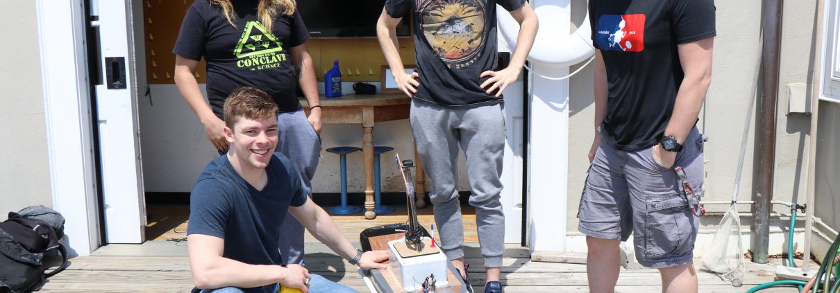 Four male students stand on the dock at the MIT Sailing Pavilion with a surface vehicle resembling a miniature boat that they developed