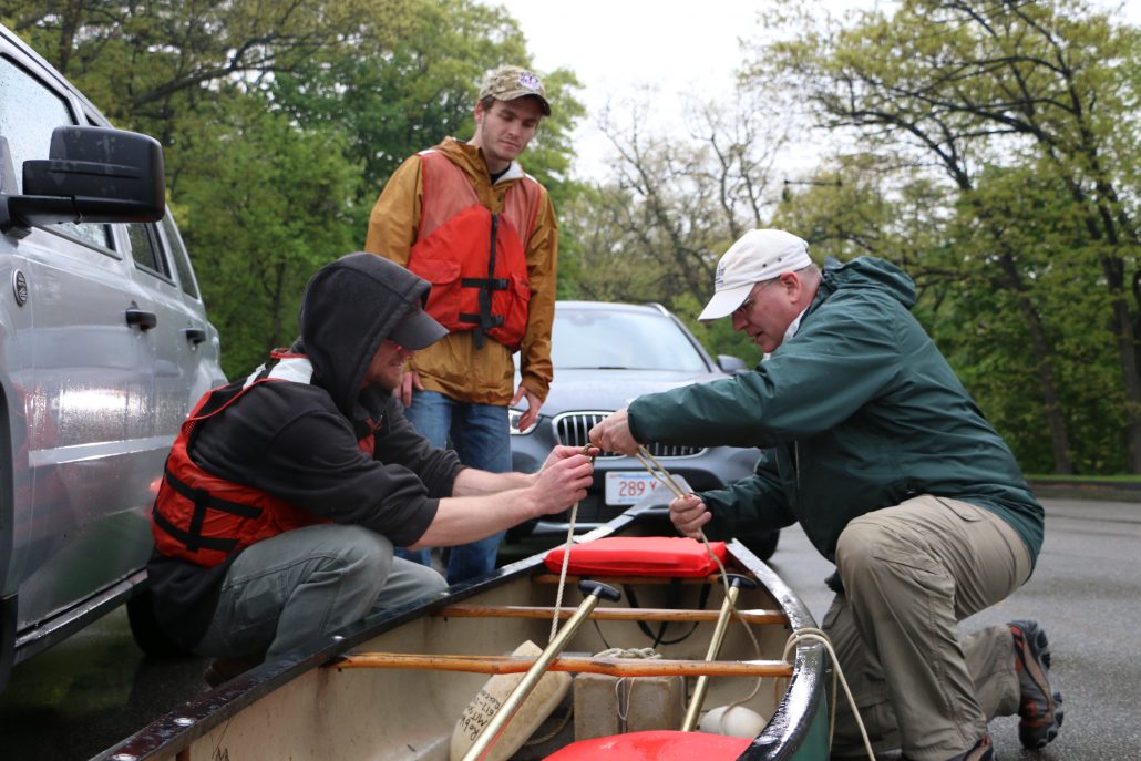 Three people work to tie ropes on a canoe in a parking lot