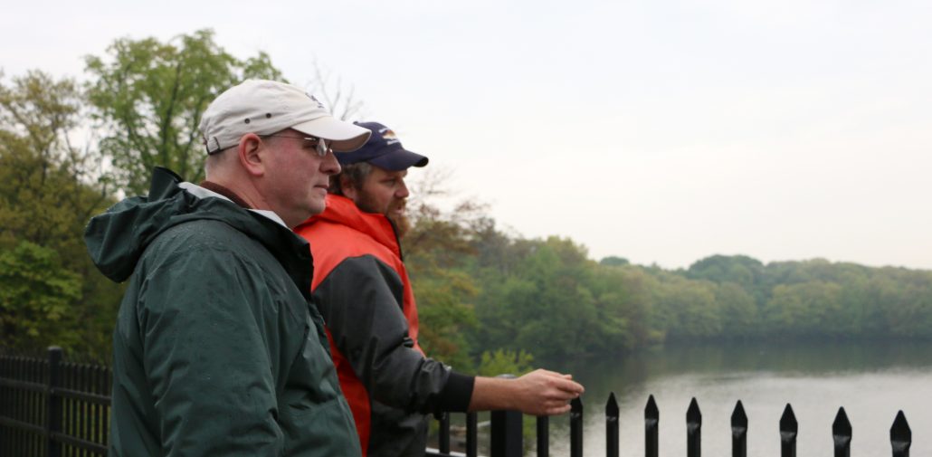 Two representatives from MIT Sea Grant and the MA DMF wearing baseball caps and rain gear look out over the Mystic Lakes