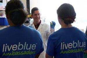 Two students from behind listen to water quality talk by a researcher holding a sample in a lab.