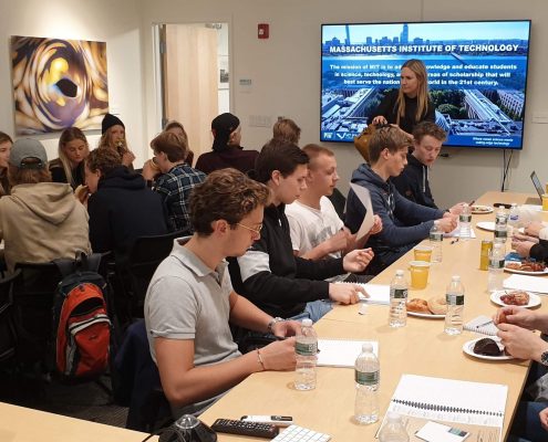 A group of students sitting for a presentation with a female speaker in front of a screen displaying a picture of the MIT campus